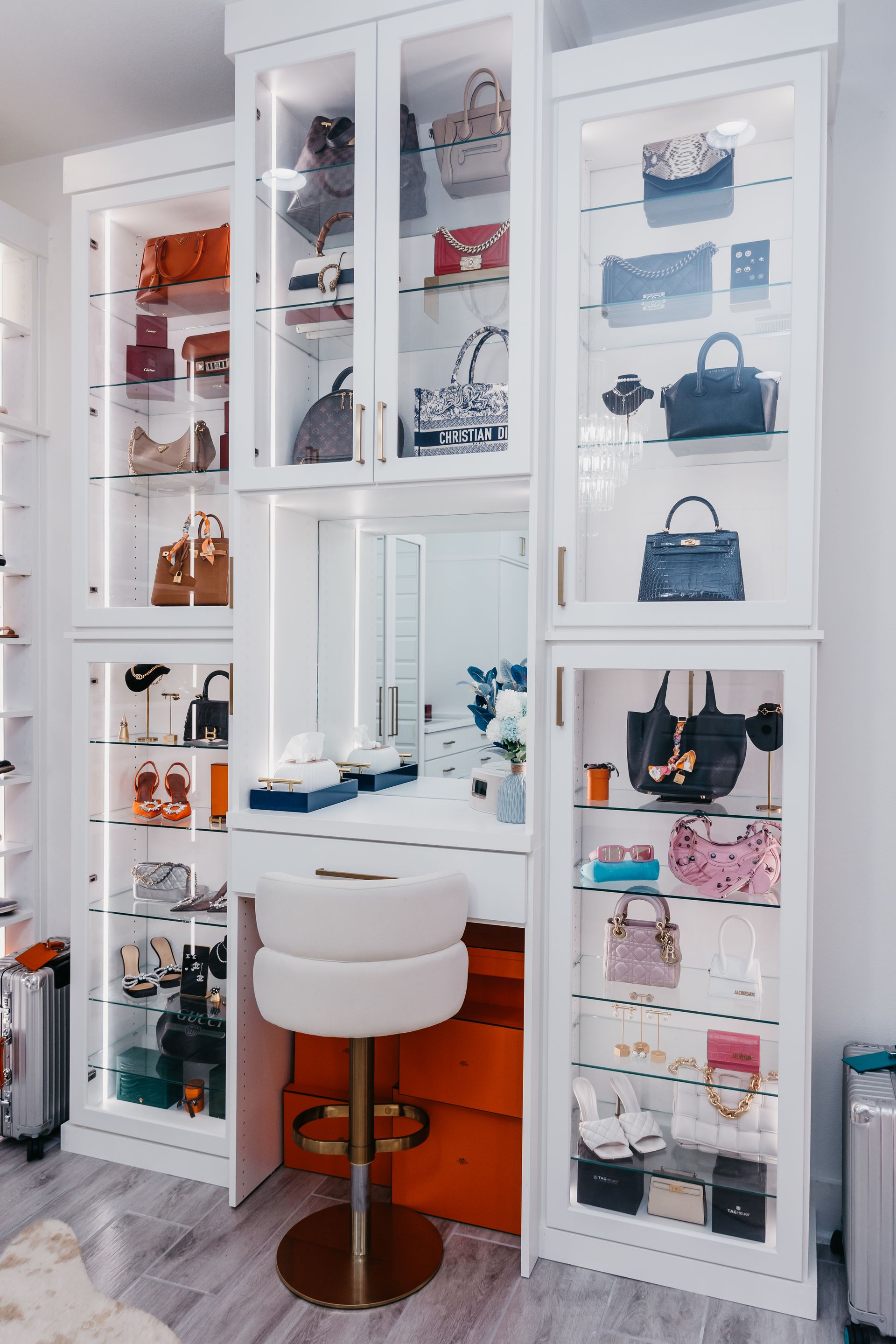 A luxurious white closet with illuminated glass cabinets displaying handbags and shoes around a vanity with a round, white stool.