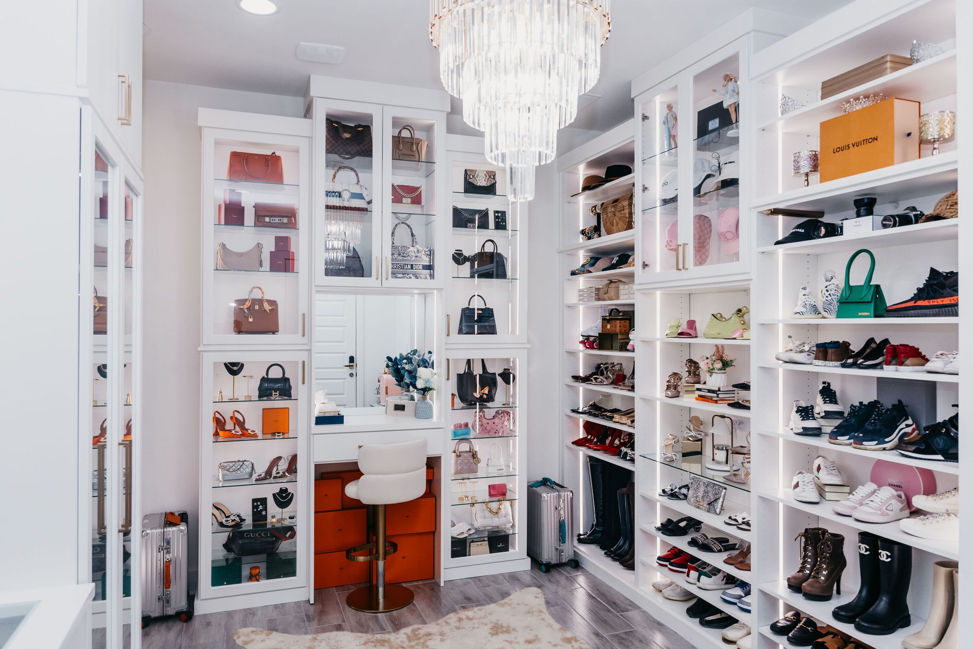 Walk-in closet with built-in shelving displaying shoes, handbags, and accessories. A vanity with a white chair sits in the center under a chandelier.