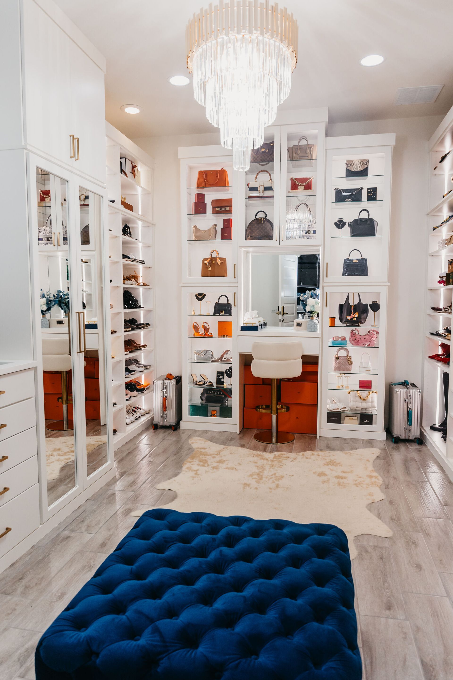 Luxurious walk-in closet with white cabinetry, glass-front shelves displaying handbags, and a vanity area. A blue ottoman sits in the foreground.