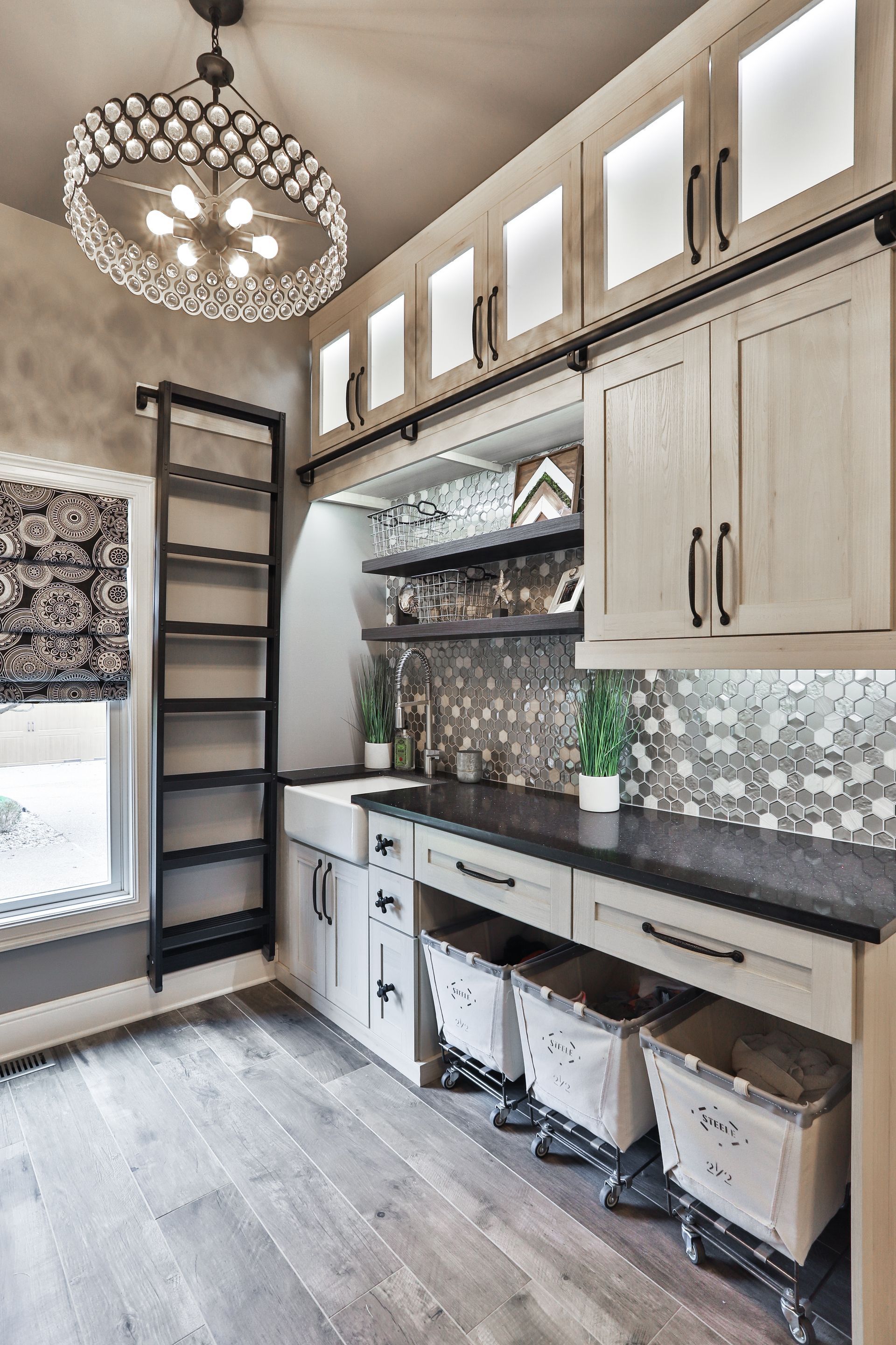 Well-lit laundry room with light wood cabinets, black countertops, a farmhouse sink, and a rolling ladder; a large circular chandelier hangs from the ceiling.
