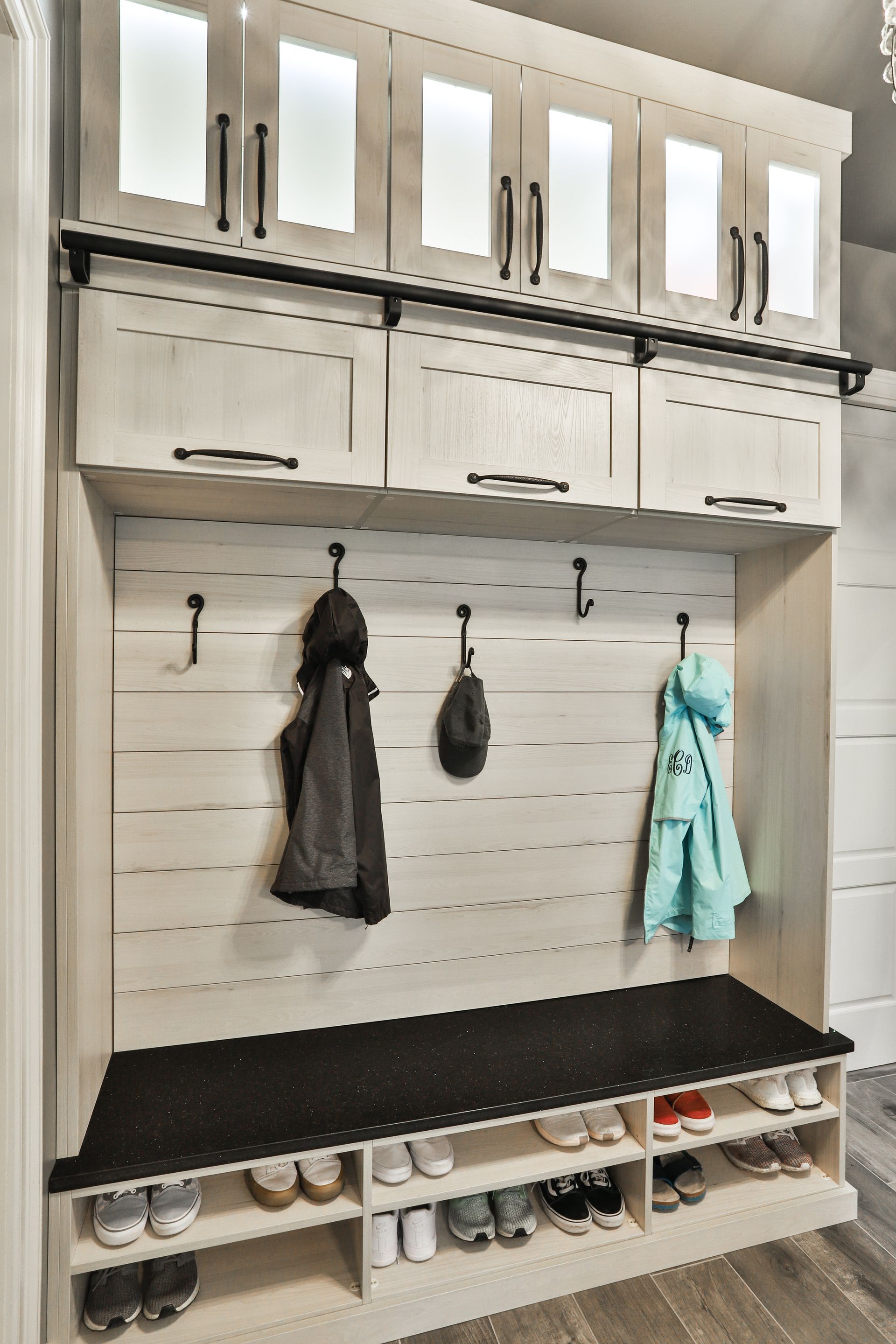 A built-in entryway storage unit with light-colored cabinets, a bench, hooks, and shoe storage. Black accents and visible clothing and a hat.
