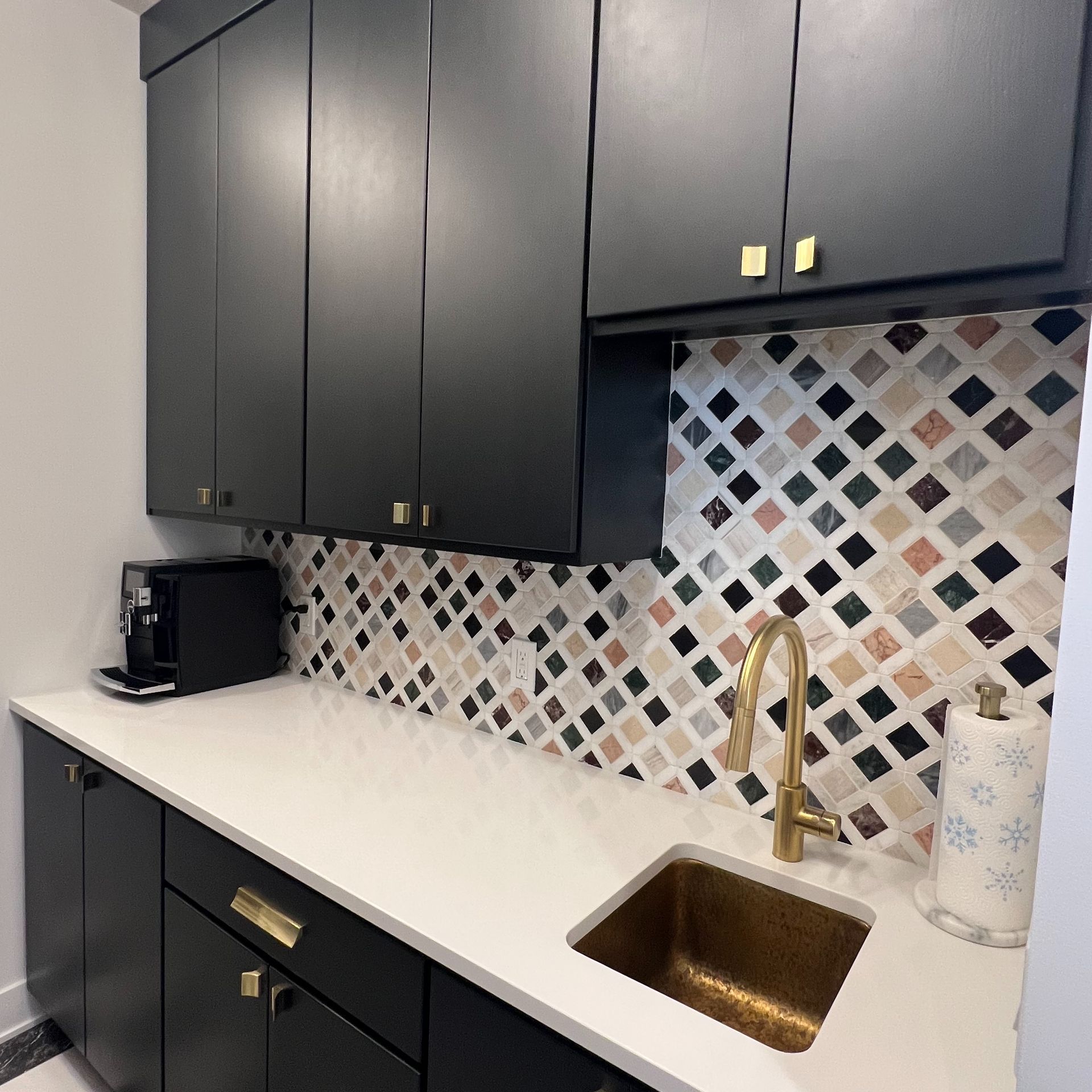 A modern kitchen counter with dark cabinets, white countertops, a brass sink and faucet, and a patterned tile backsplash.