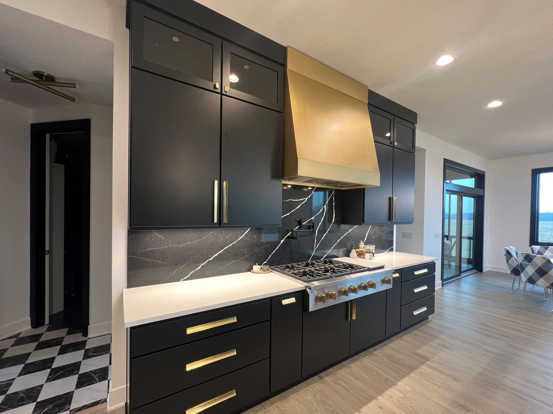 A modern kitchen featuring matte black cabinets, a gold vent hood, a black marble-style backsplash, and white countertops.