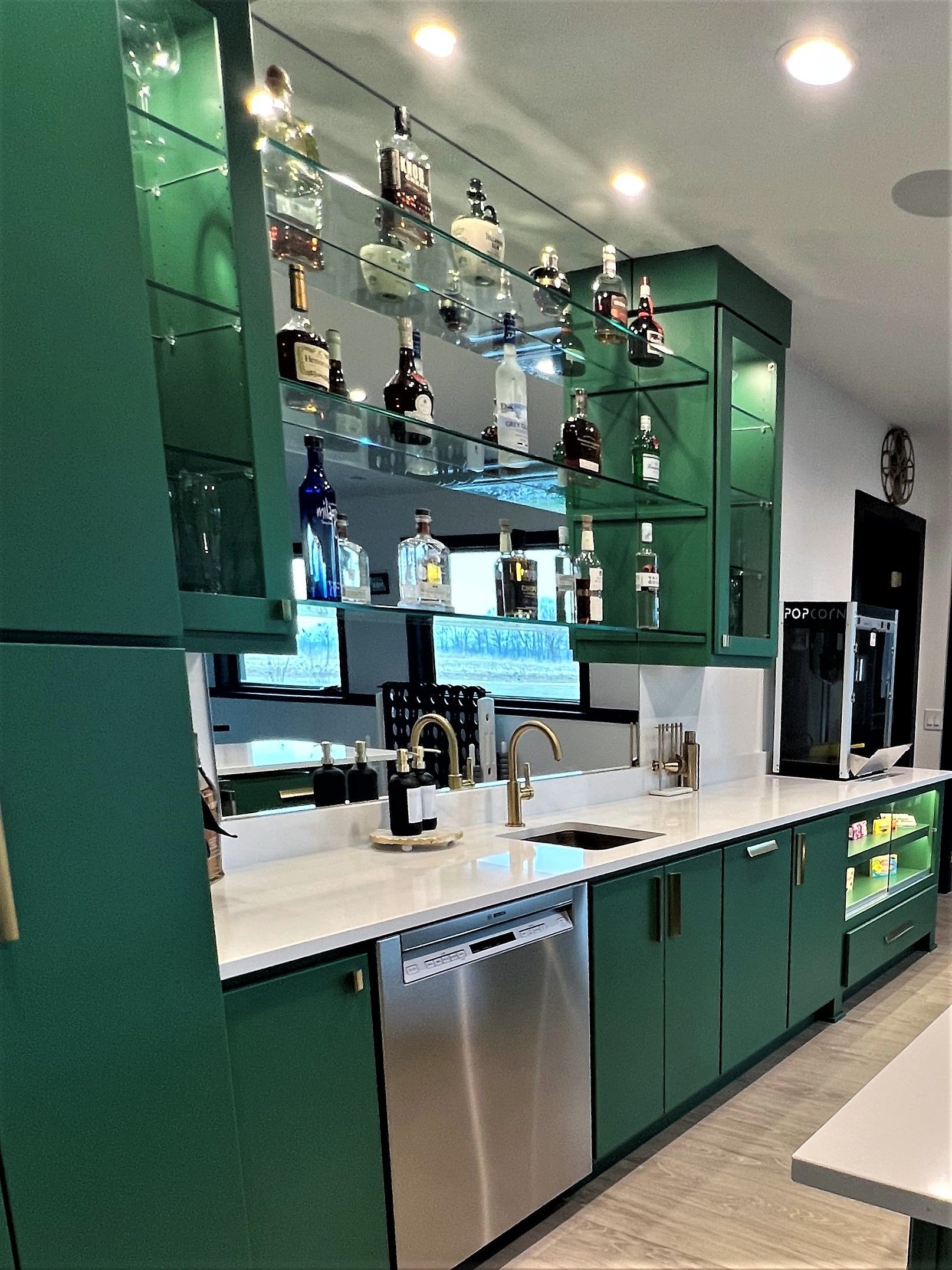 A modern home bar with dark green cabinets, white countertops, stainless steel appliances, and tiered glass bottle shelving.