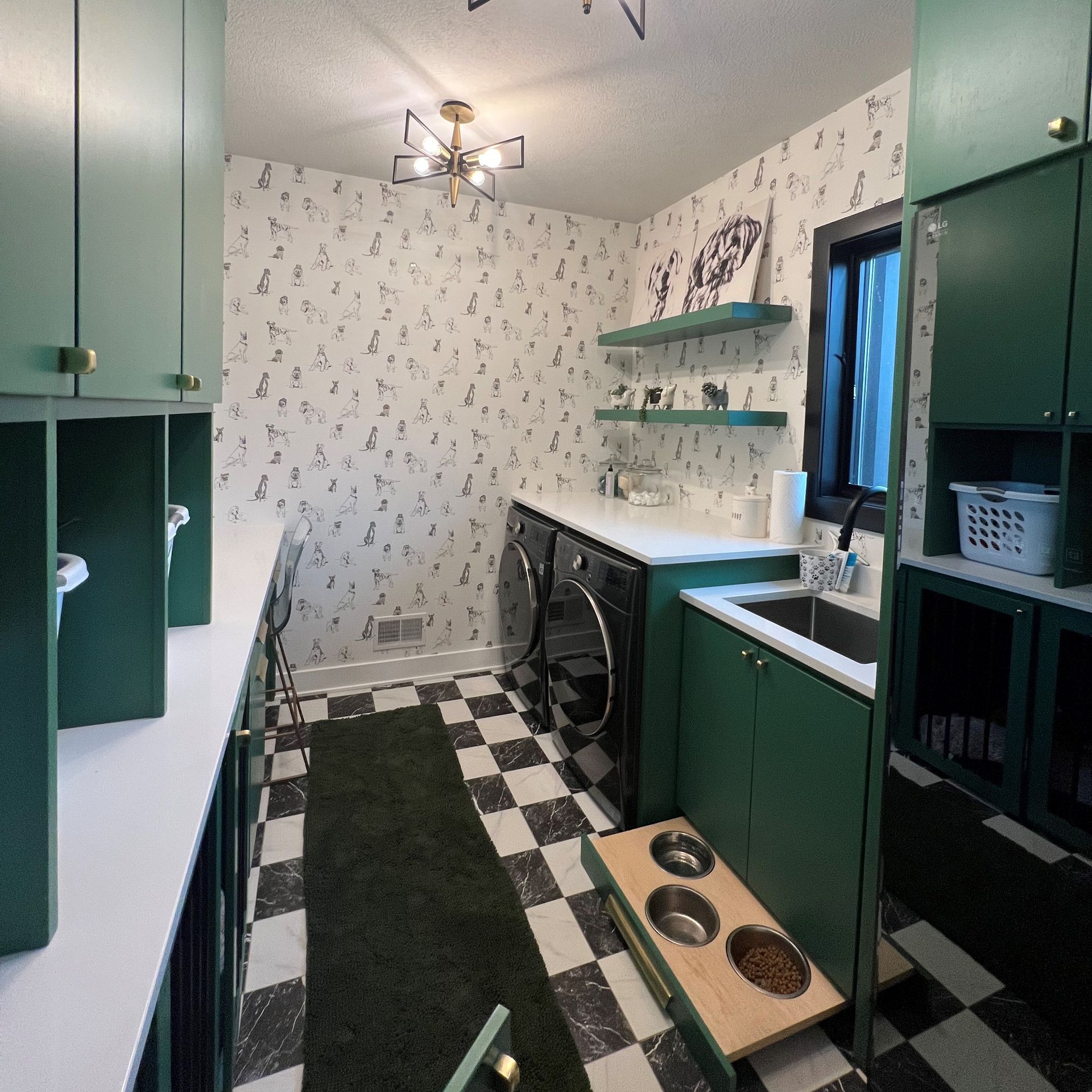 A laundry room with green cabinets, patterned wallpaper, a checkered floor, two washers, and a pull-out pet feeding drawer.
