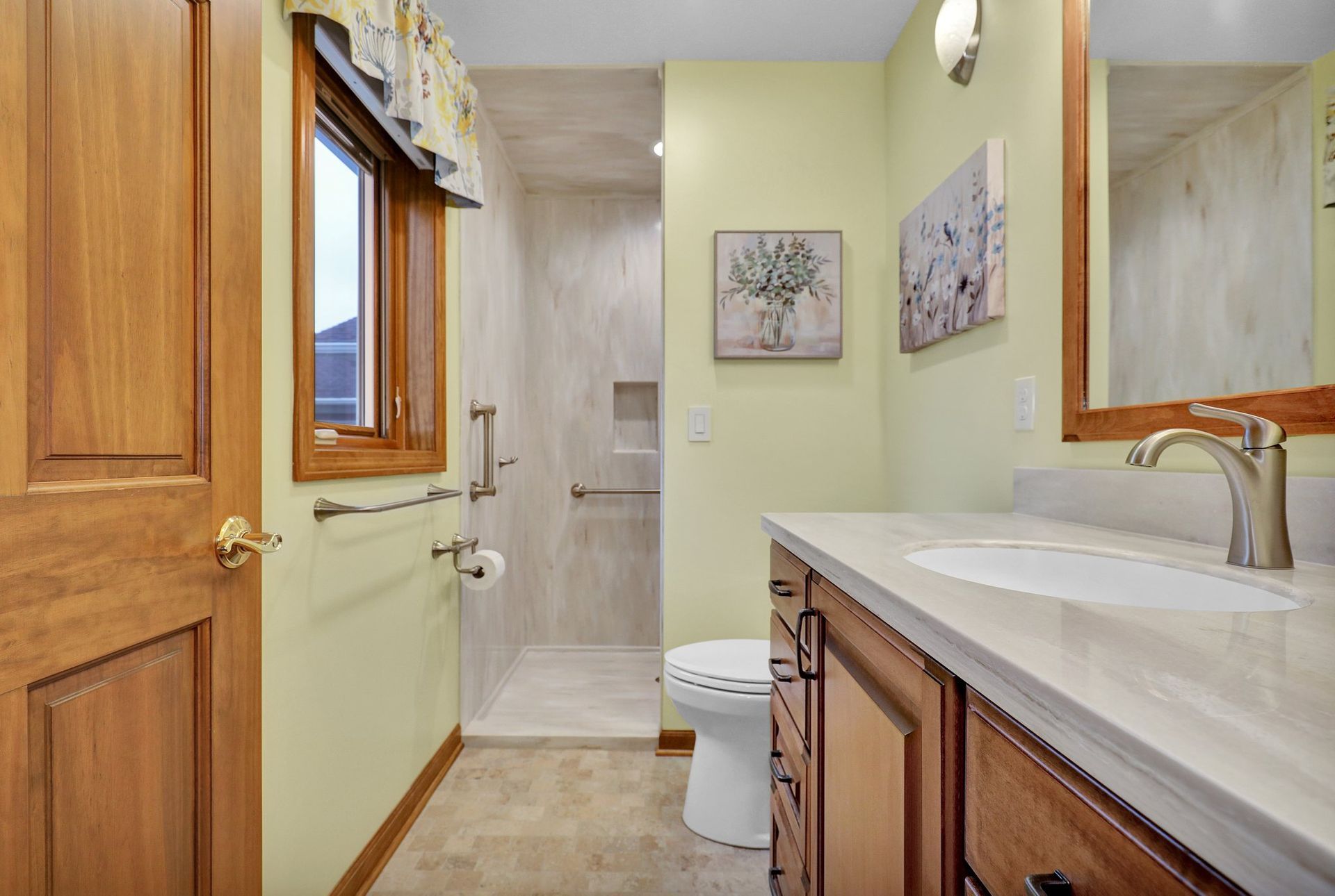 A bathroom with a wooden vanity, a beige countertop sink, a white toilet, and a tiled walk-in shower with safety bars.