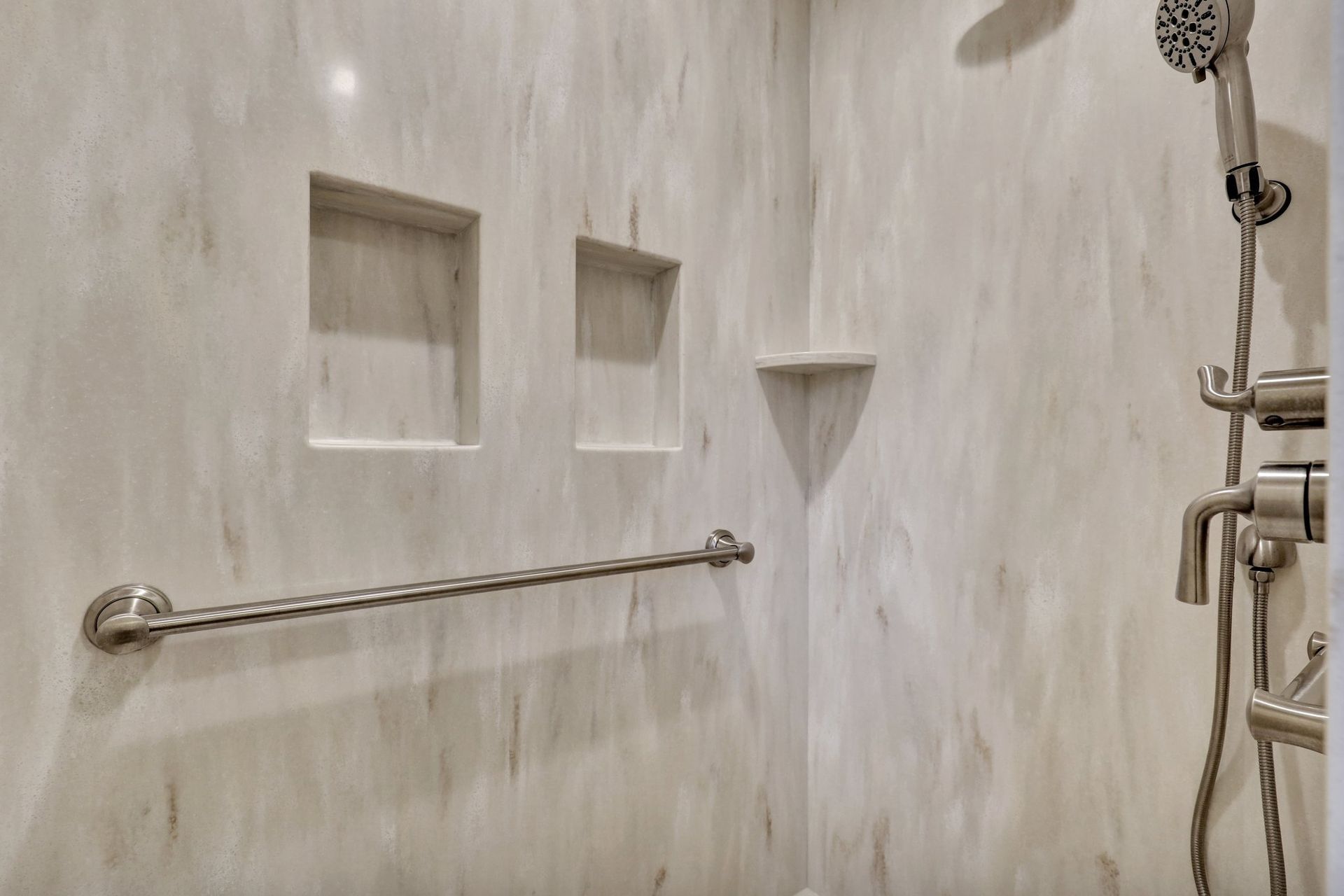 Shower stall with light marble-patterned walls, a horizontal grab bar, two recessed wall niches, and a handheld shower head.