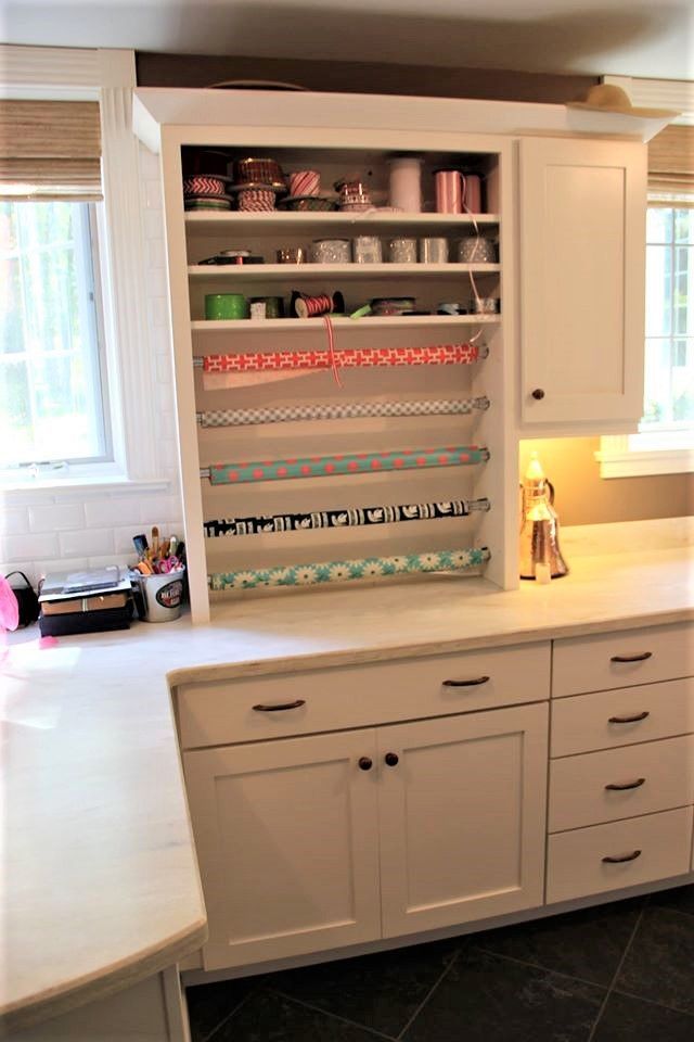 White wooden craft station featuring built-in shelving for wrapping paper rolls and drawers for storage in a bright room.