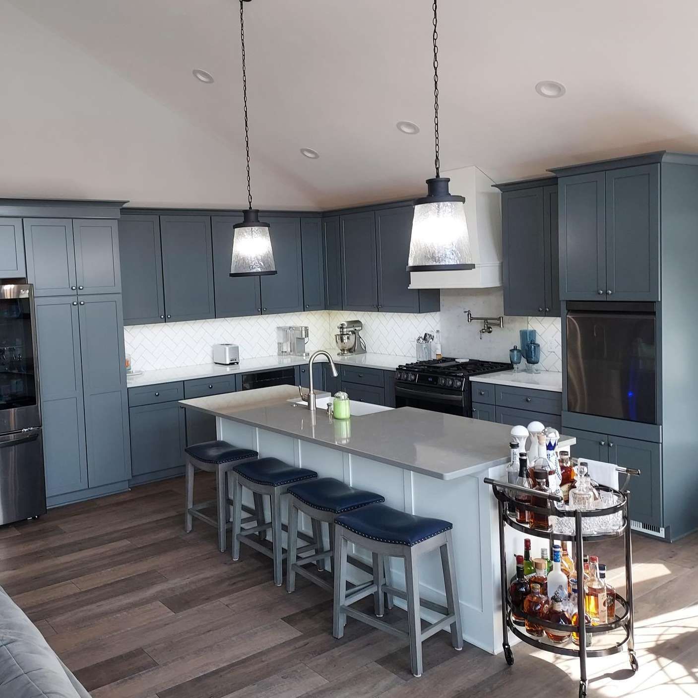 Modern kitchen with dark grey cabinets, a central white island with seating, pendant lights, and a rolling drink cart.
