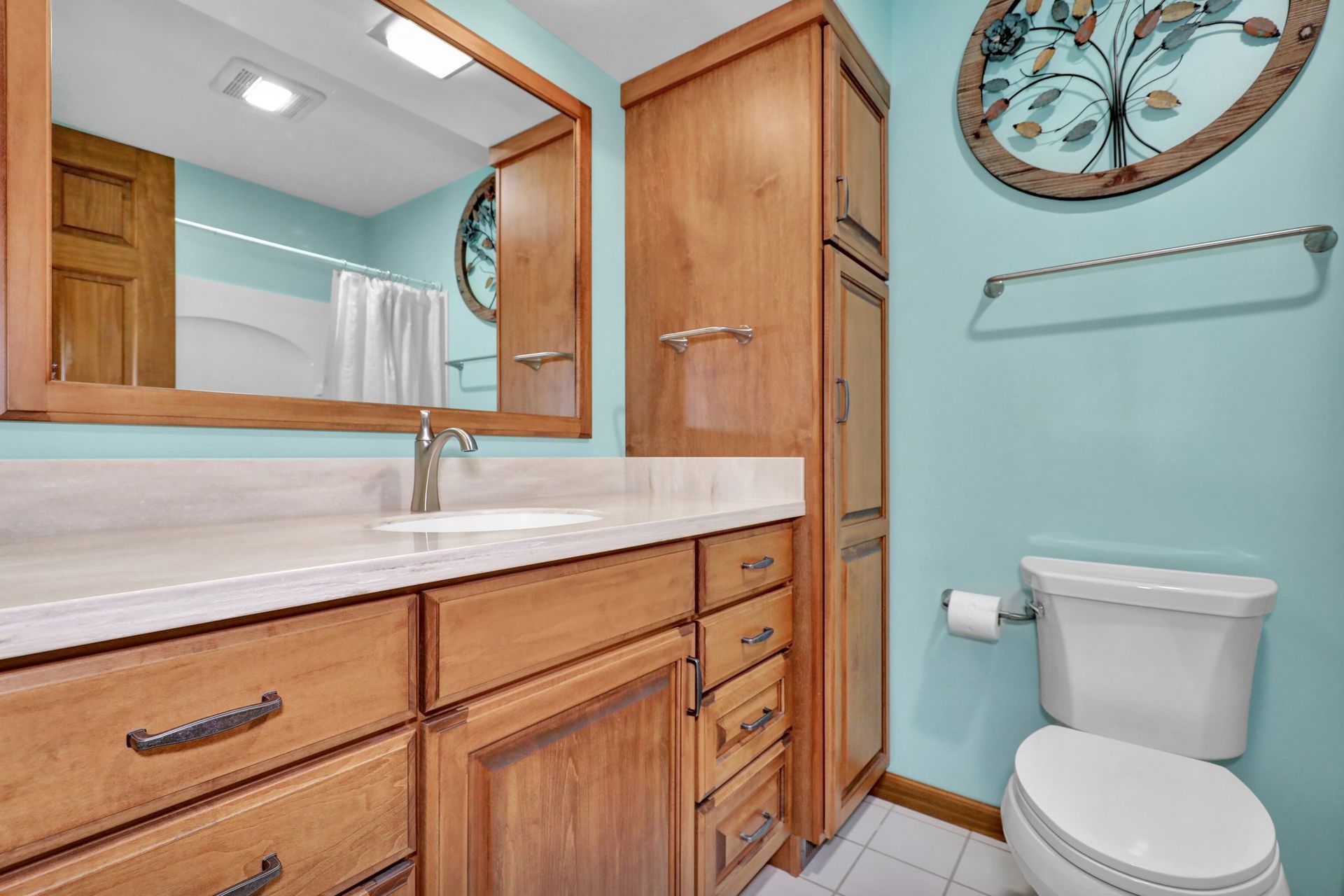 A light blue bathroom with a wooden vanity, matching tall cabinet, a large mirror, and a white toilet.