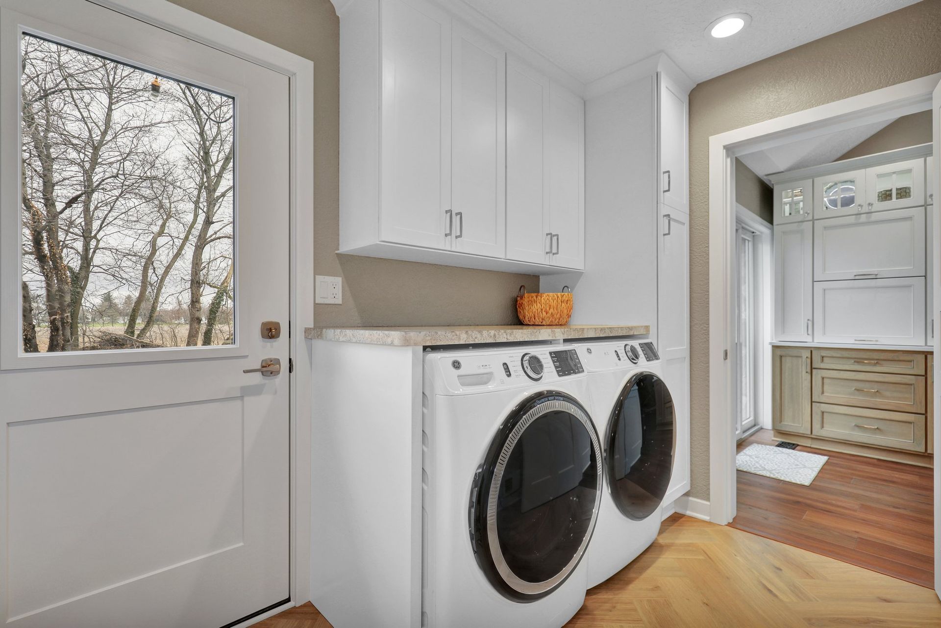 A laundry room with a white washer and dryer, light wood floor, white cabinetry, and an exterior door looking outside.