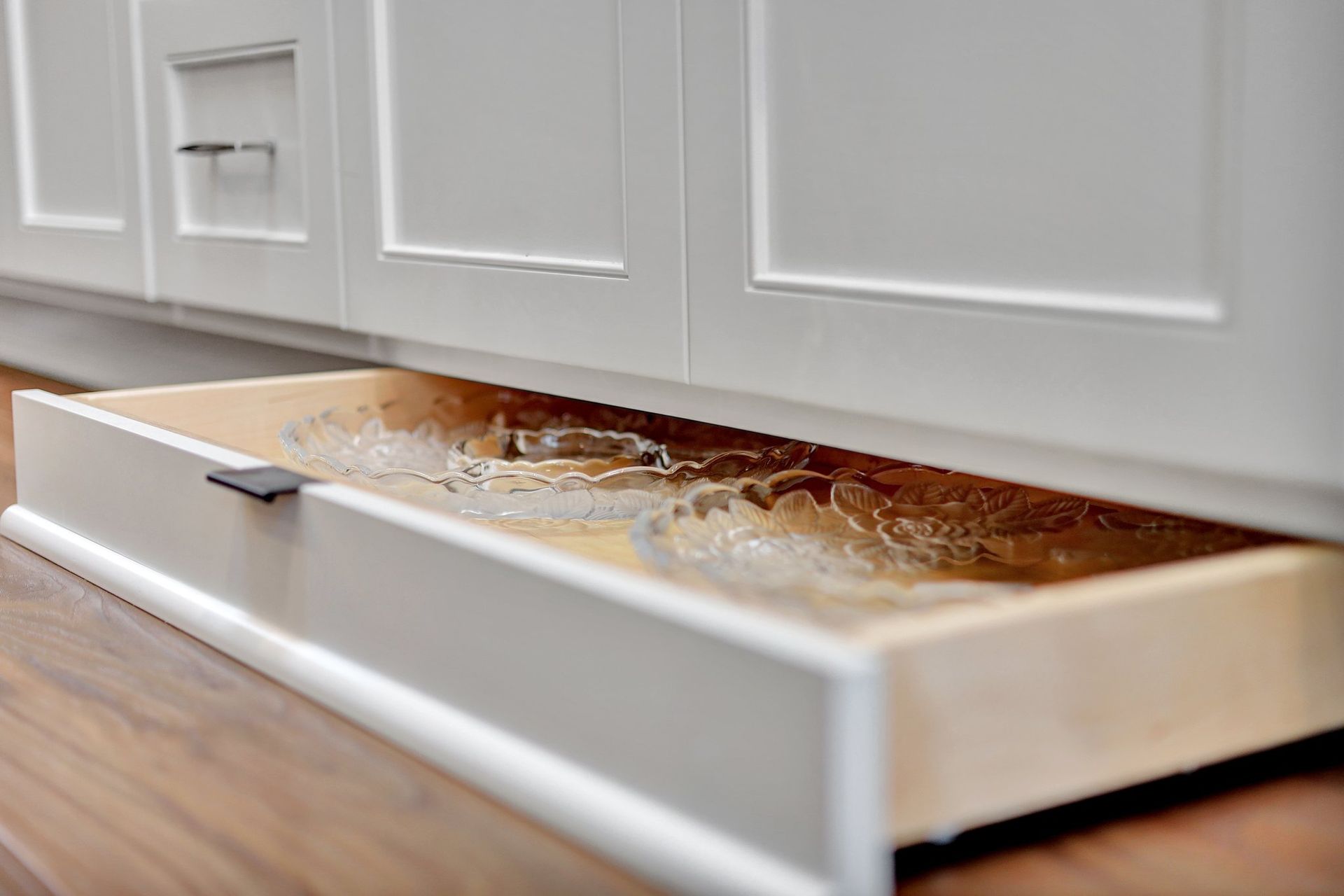A low-profile white drawer is pulled open on a wood floor, revealing two clear, decorative glass bowls inside.