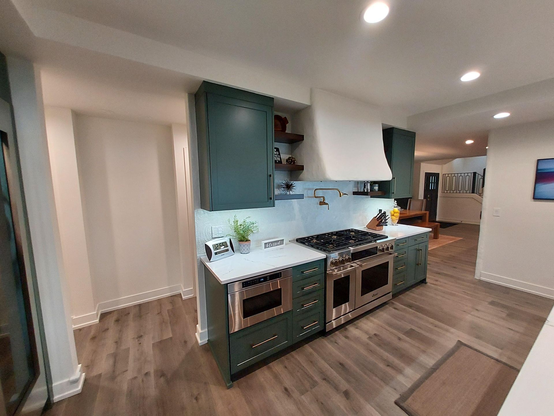A kitchen with dark green cabinets, white countertops, a large custom range hood, and stainless steel appliances.
