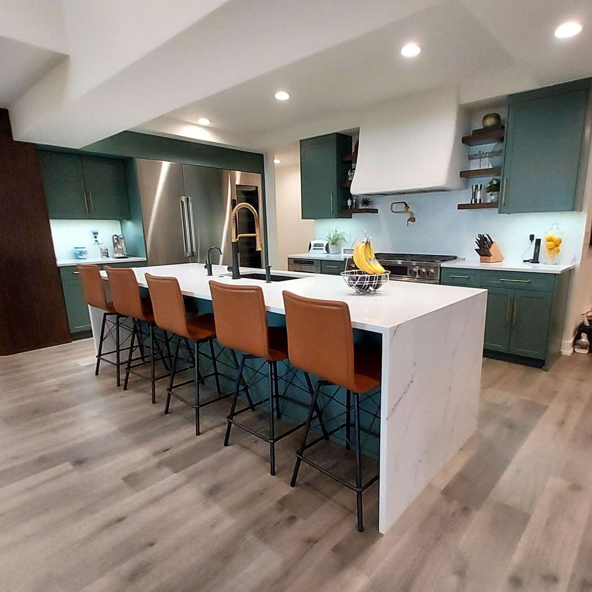 A modern kitchen with deep green cabinets, white marble countertops, and four brown leather bar stools on light wood floors.