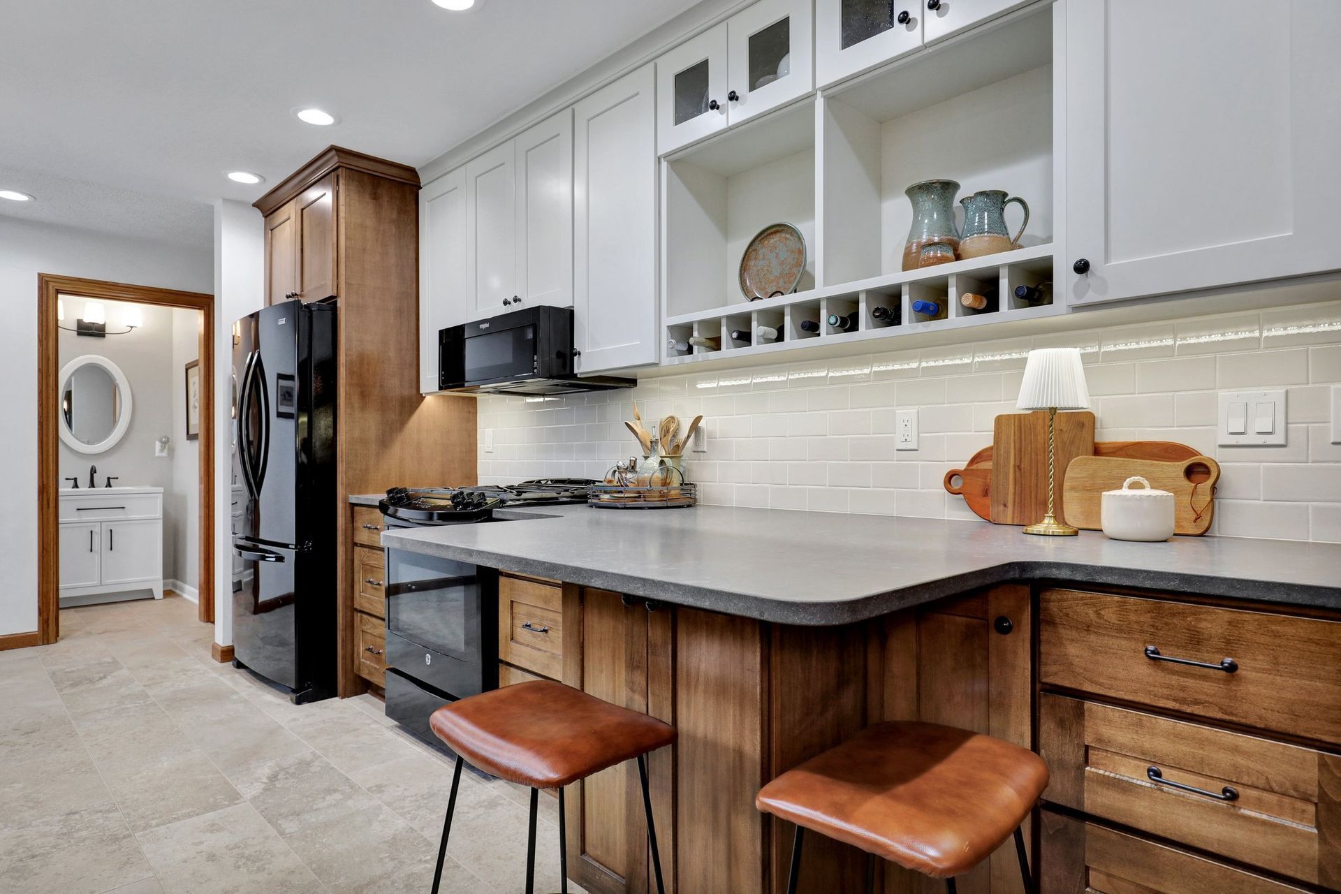 A kitchen with a gray countertop, wood cabinets, white upper cabinets, a black refrigerator, and two leather stools.
