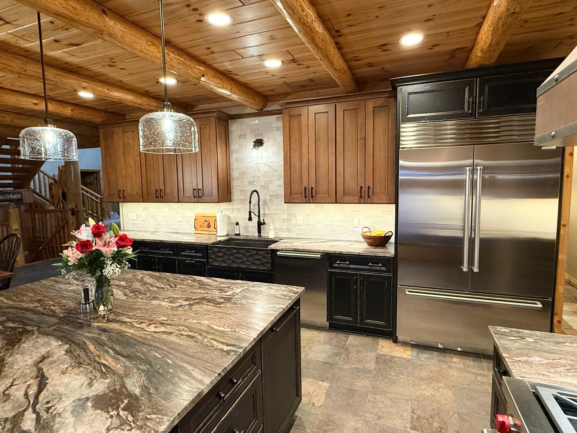 A rustic kitchen features wood-paneled ceilings, stone countertops, a large stainless steel refrigerator, and dark cabinets.