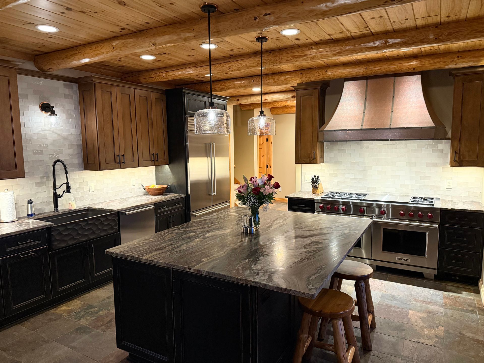 A rustic kitchen with dark cabinetry, a large stone island, log ceiling beams, and a stainless steel stove with a hood.