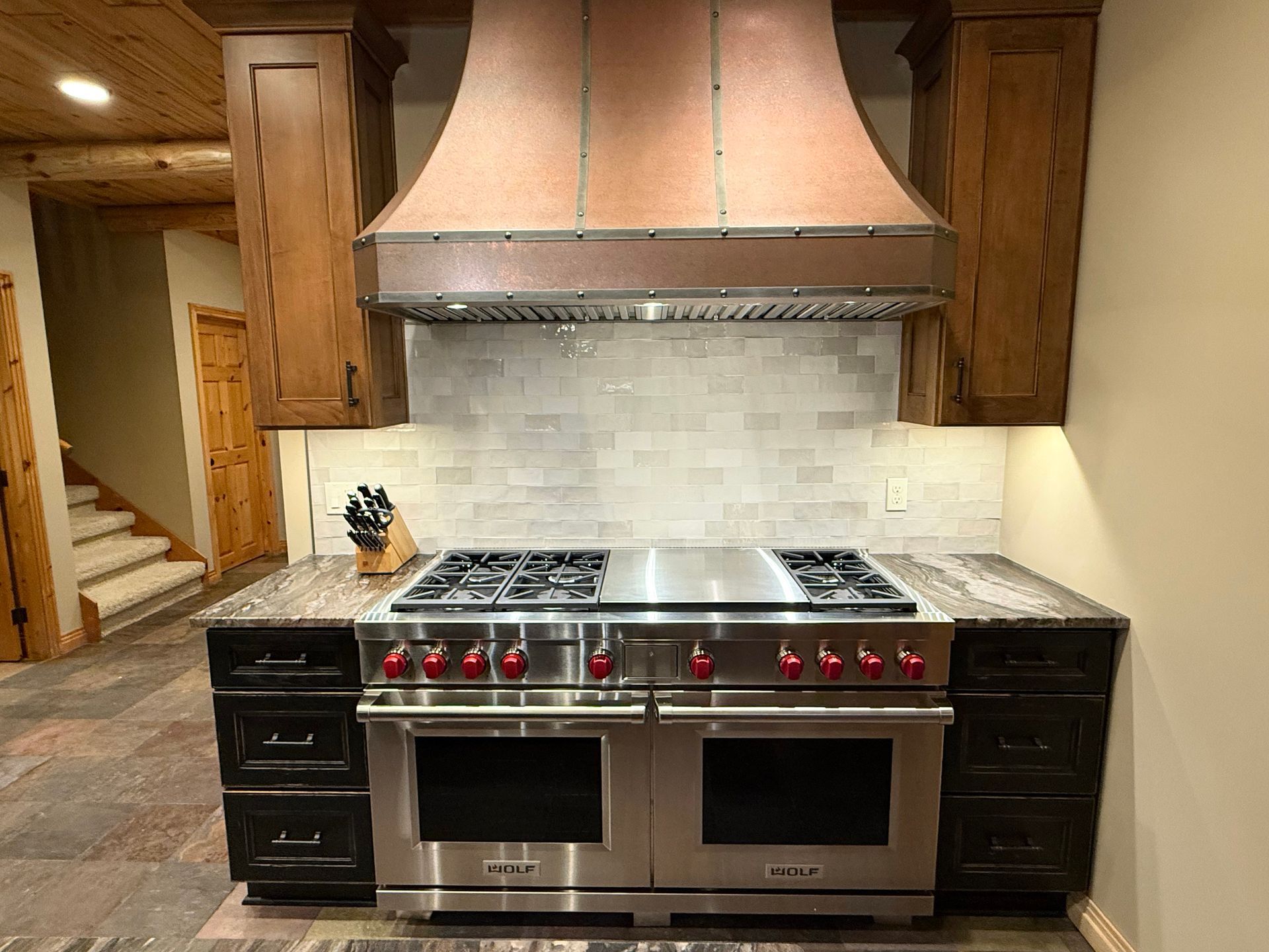 A large professional stainless steel range with red knobs sits under a copper range hood between wood cabinets.