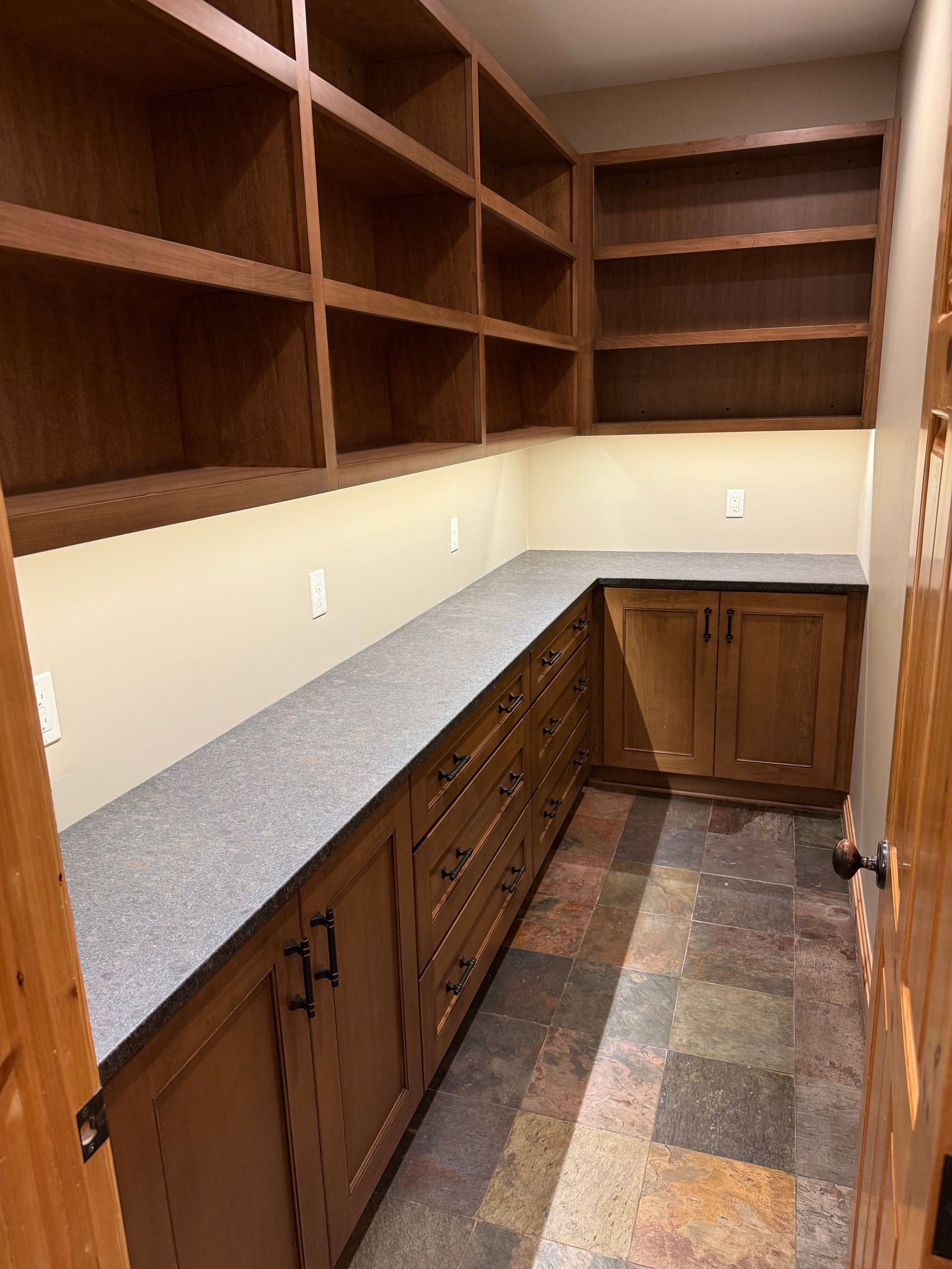 A pantry with wooden cabinets, gray countertops, open upper shelving, and tiled flooring.