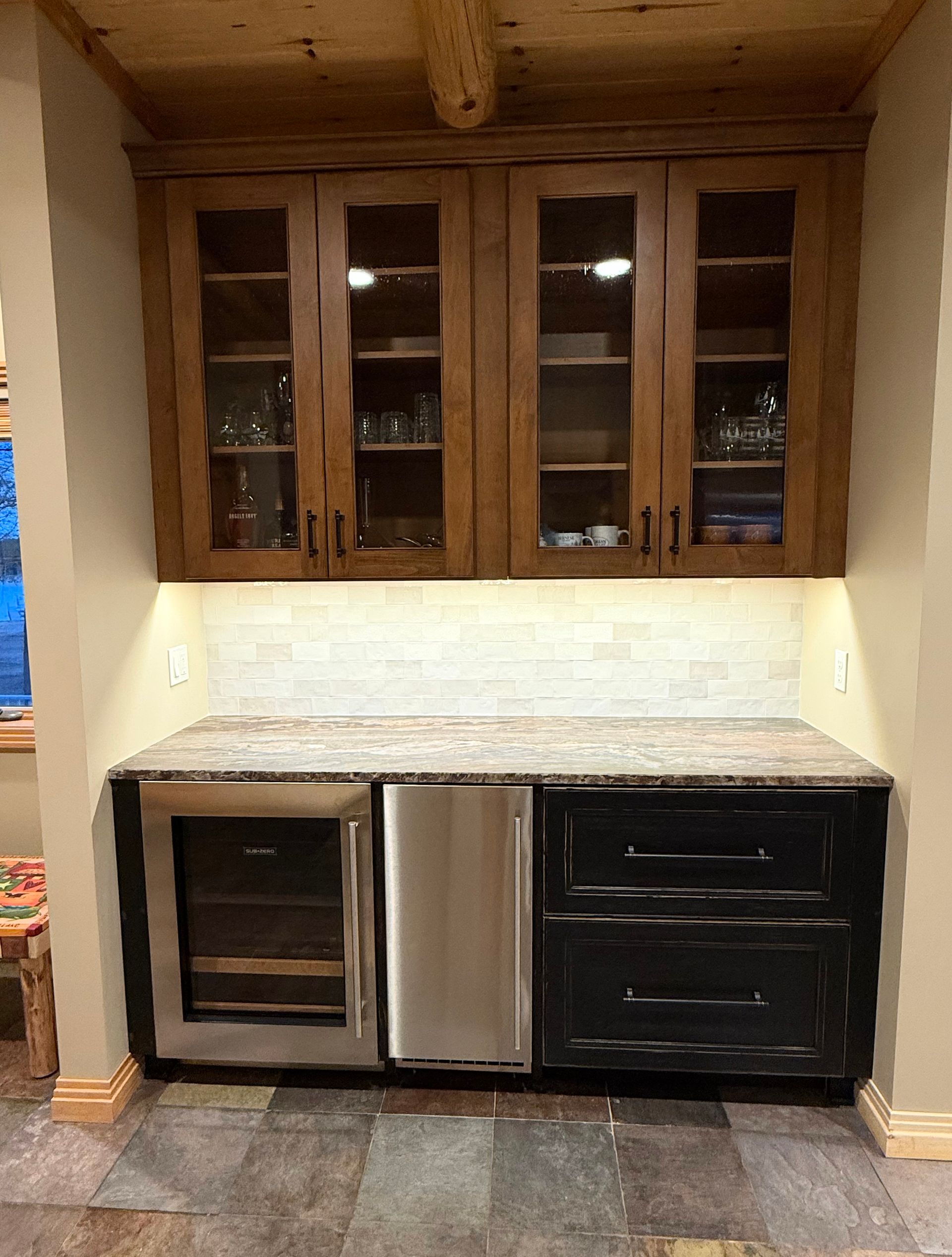 A kitchen beverage station with wood-framed glass cabinets above a counter, a wine fridge, ice maker, and black drawers.