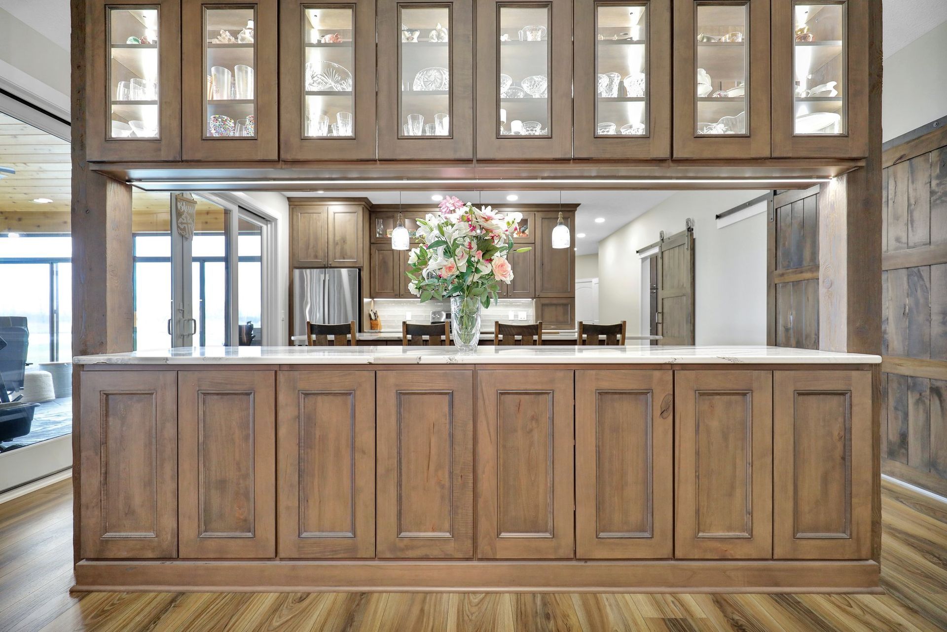 A kitchen island with warm wood cabinetry and a marble top, featuring glass-front cabinets above an open serving pass.
