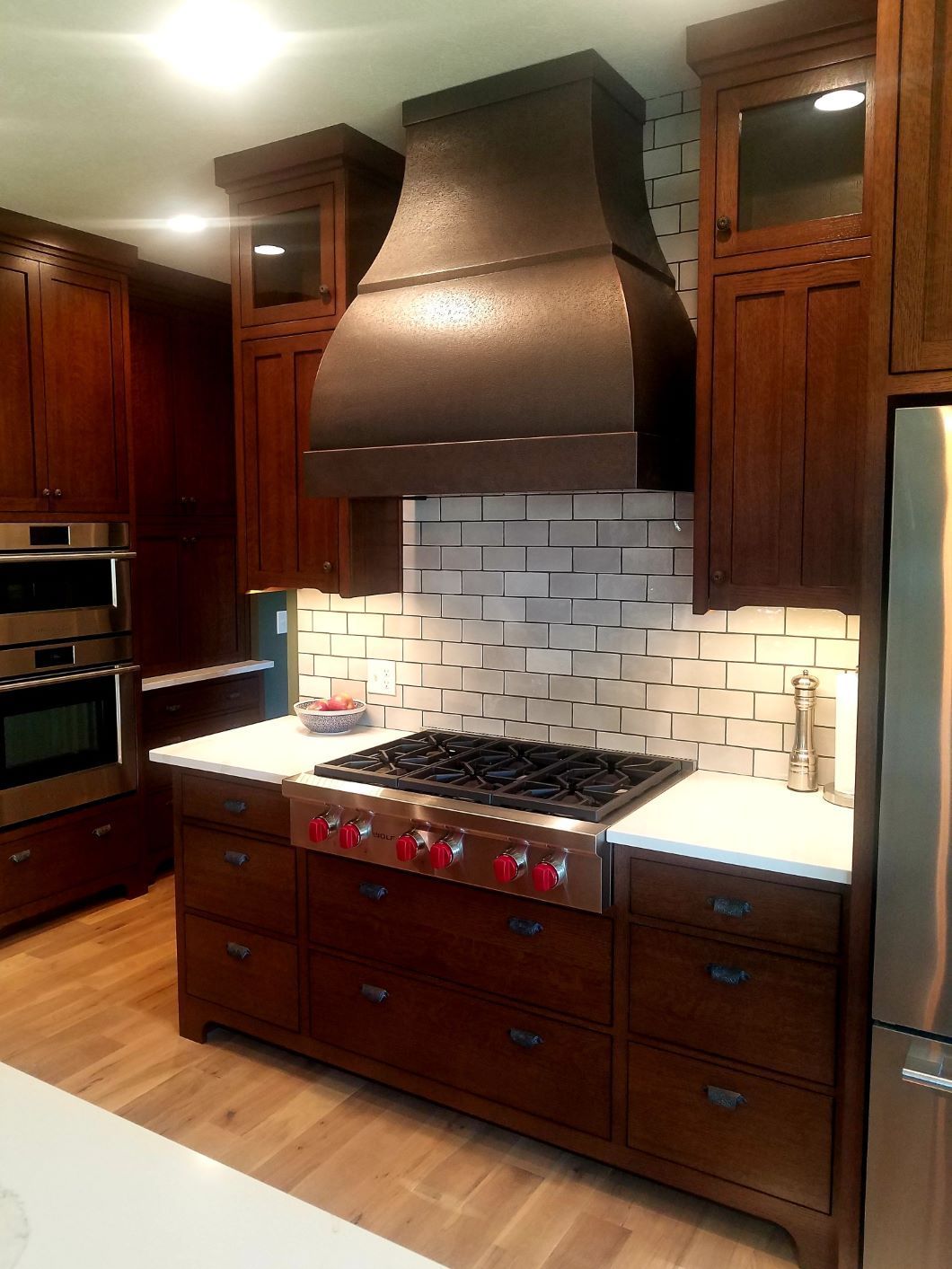 A kitchen stove area with dark wood cabinets, a large copper vent hood, white tile backsplash, and a stainless gas range.