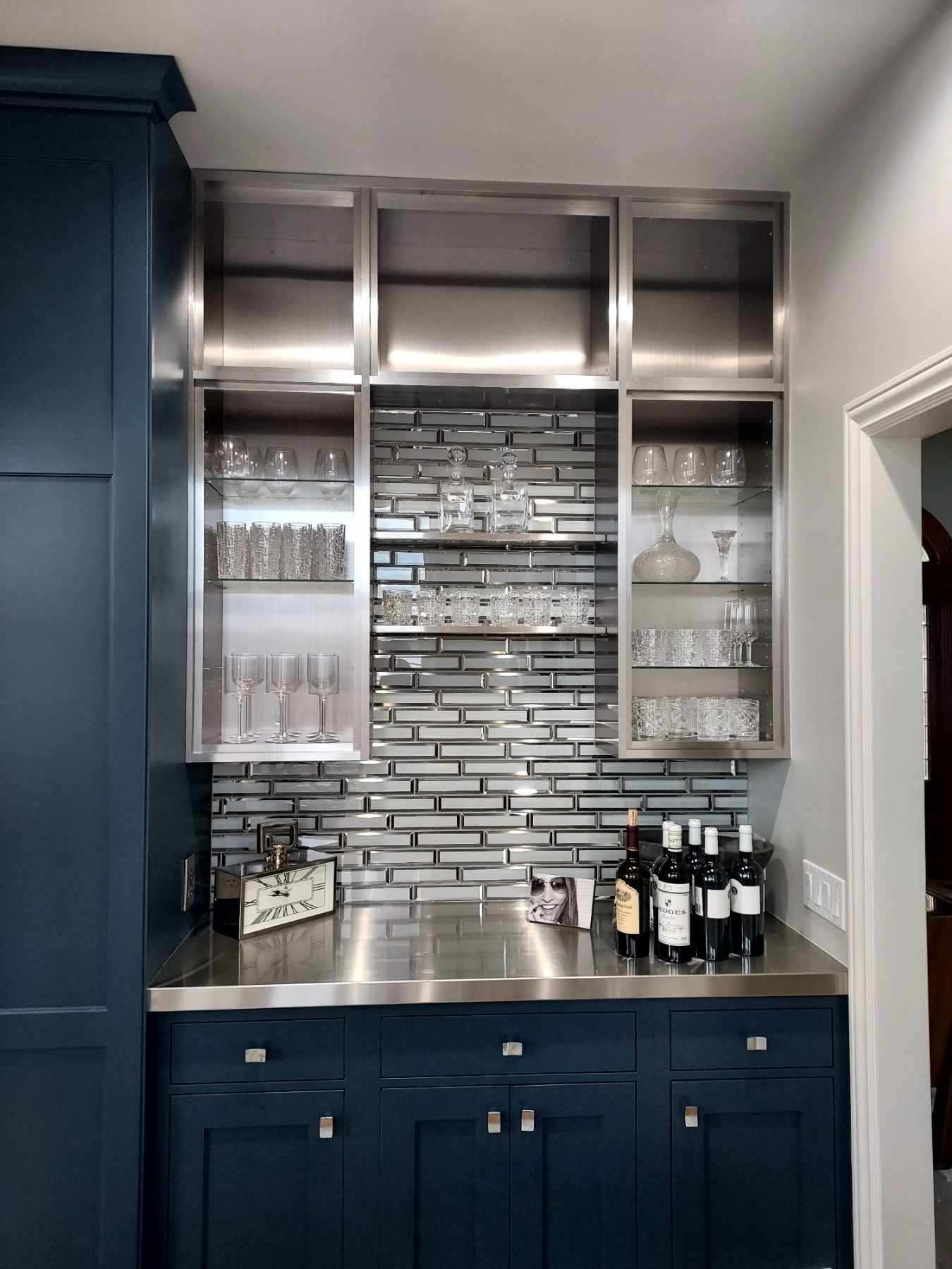 A dark blue wet bar with metal shelves, glass cabinets, and a reflective brick-patterned backsplash.