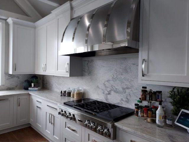 A modern kitchen featuring white cabinets, a large stainless steel range hood, and a marble-patterned backsplash.