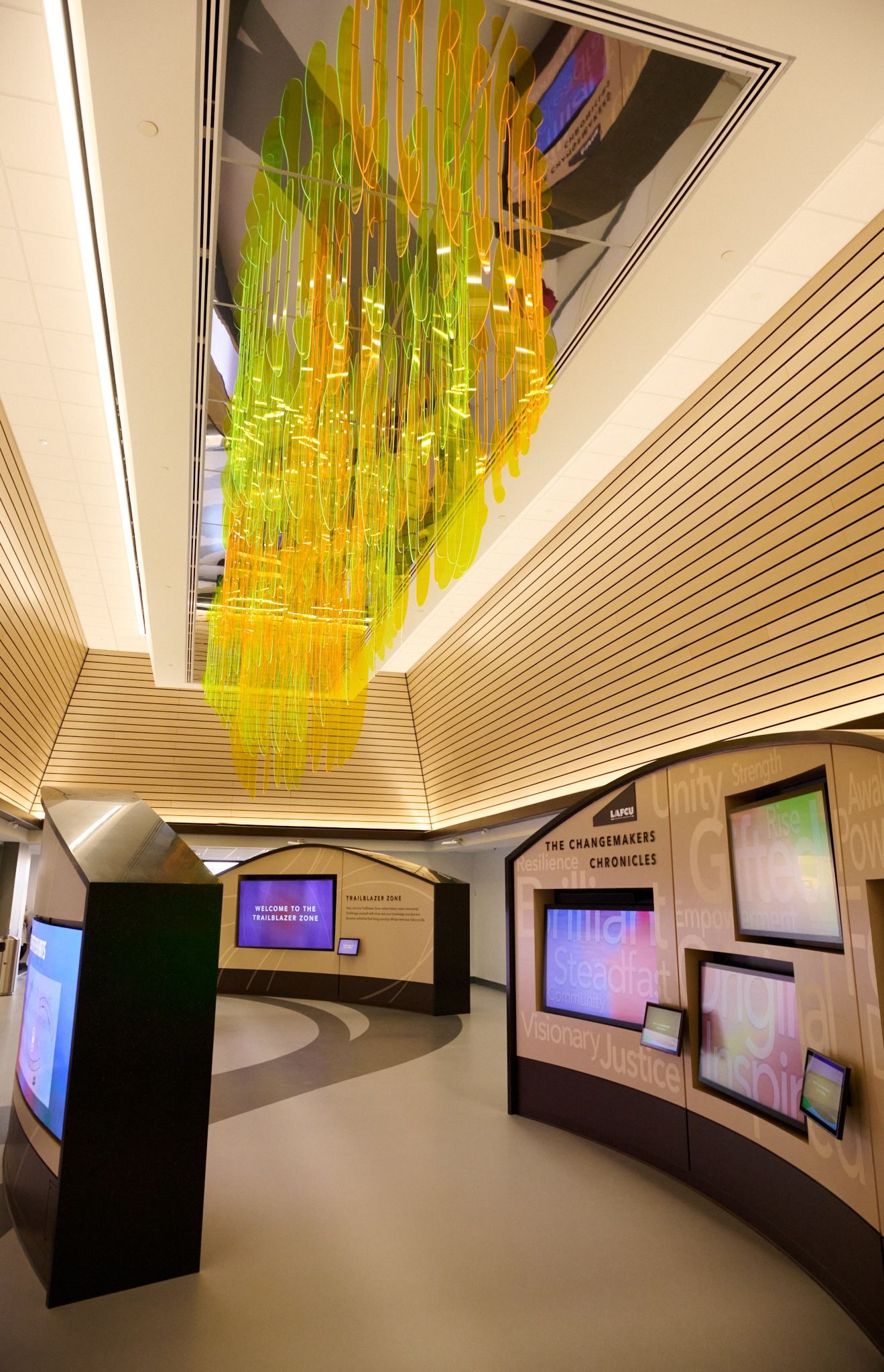A modern lobby with a large yellow glass sculpture hanging from the ceiling, wood-paneled walls, and digital kiosks.