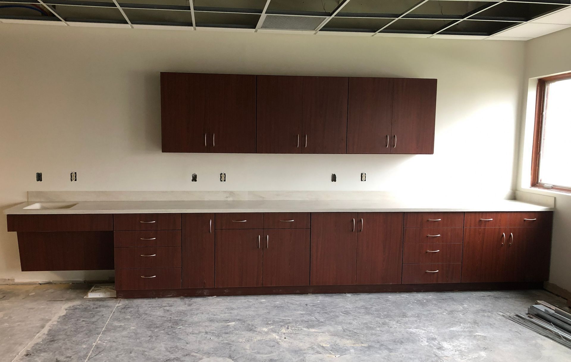 A wall of dark wood cabinets with white countertops, including a wheelchair-accessible sink area in a room under renovation.