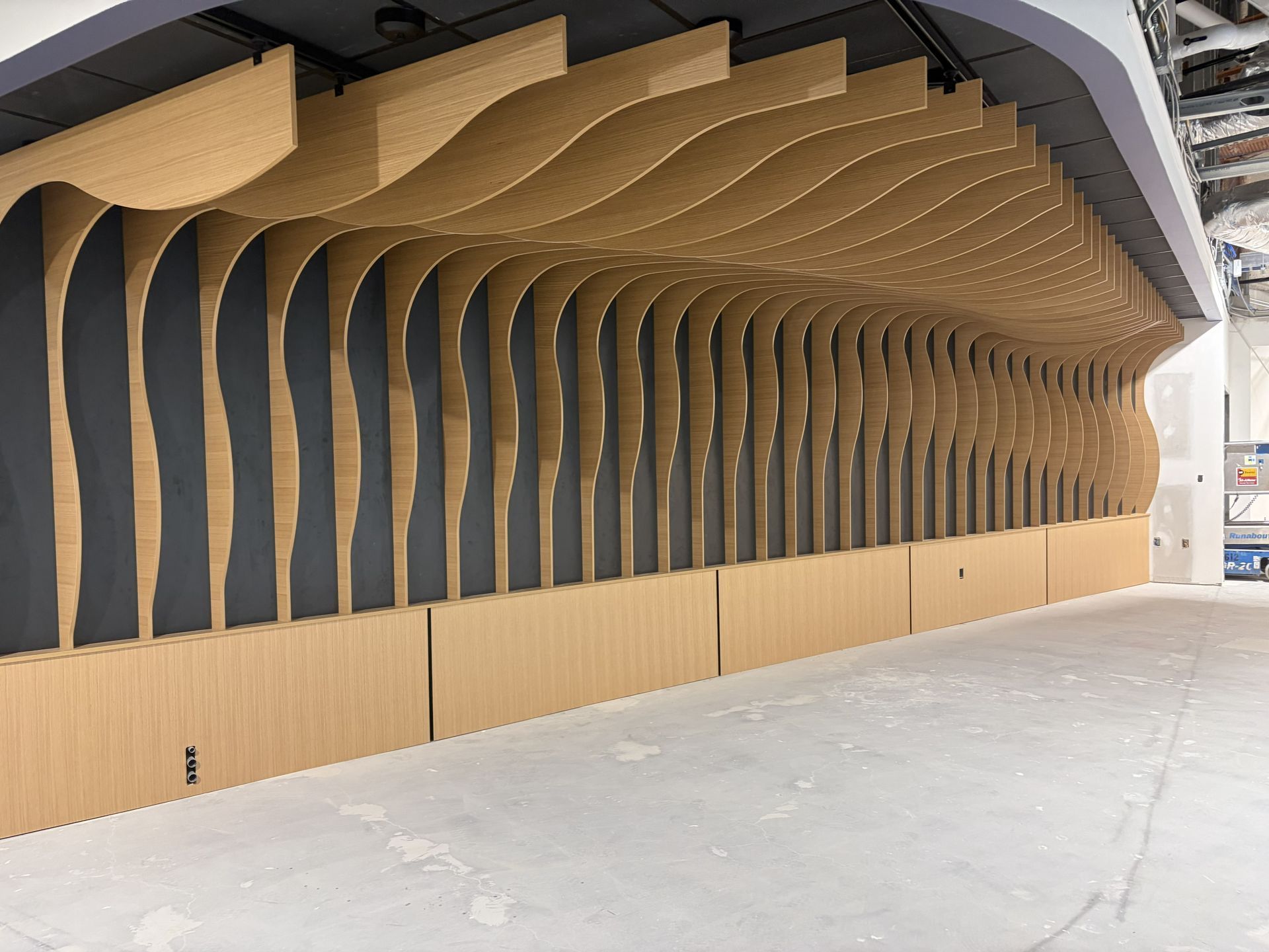 A modern wall installation featuring repetitive, wavy light-wood slats against a dark backdrop in a minimalist space.