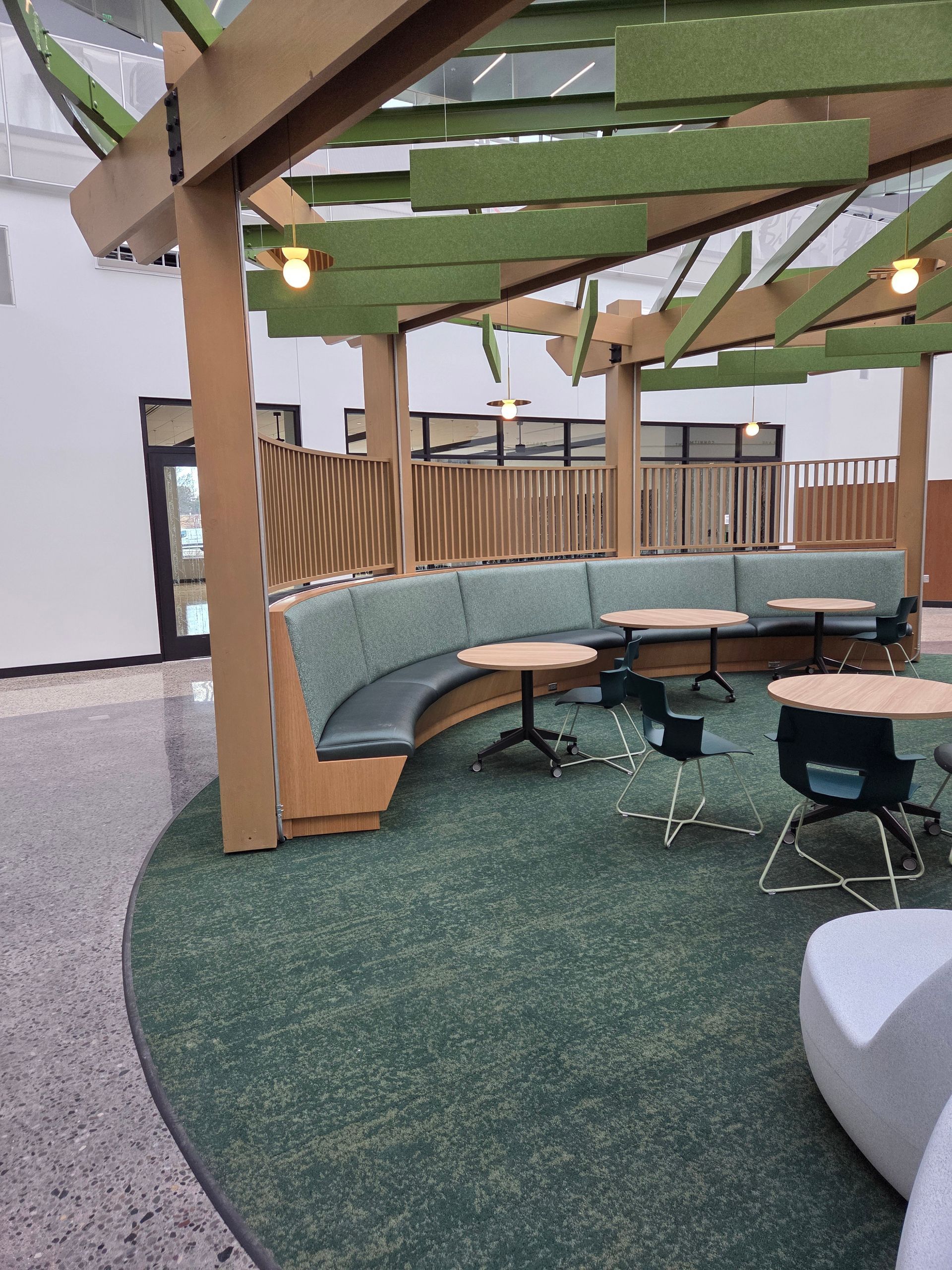 Modern lounge area with a curved teal booth, round wooden tables, and green hanging acoustic ceiling baffles.