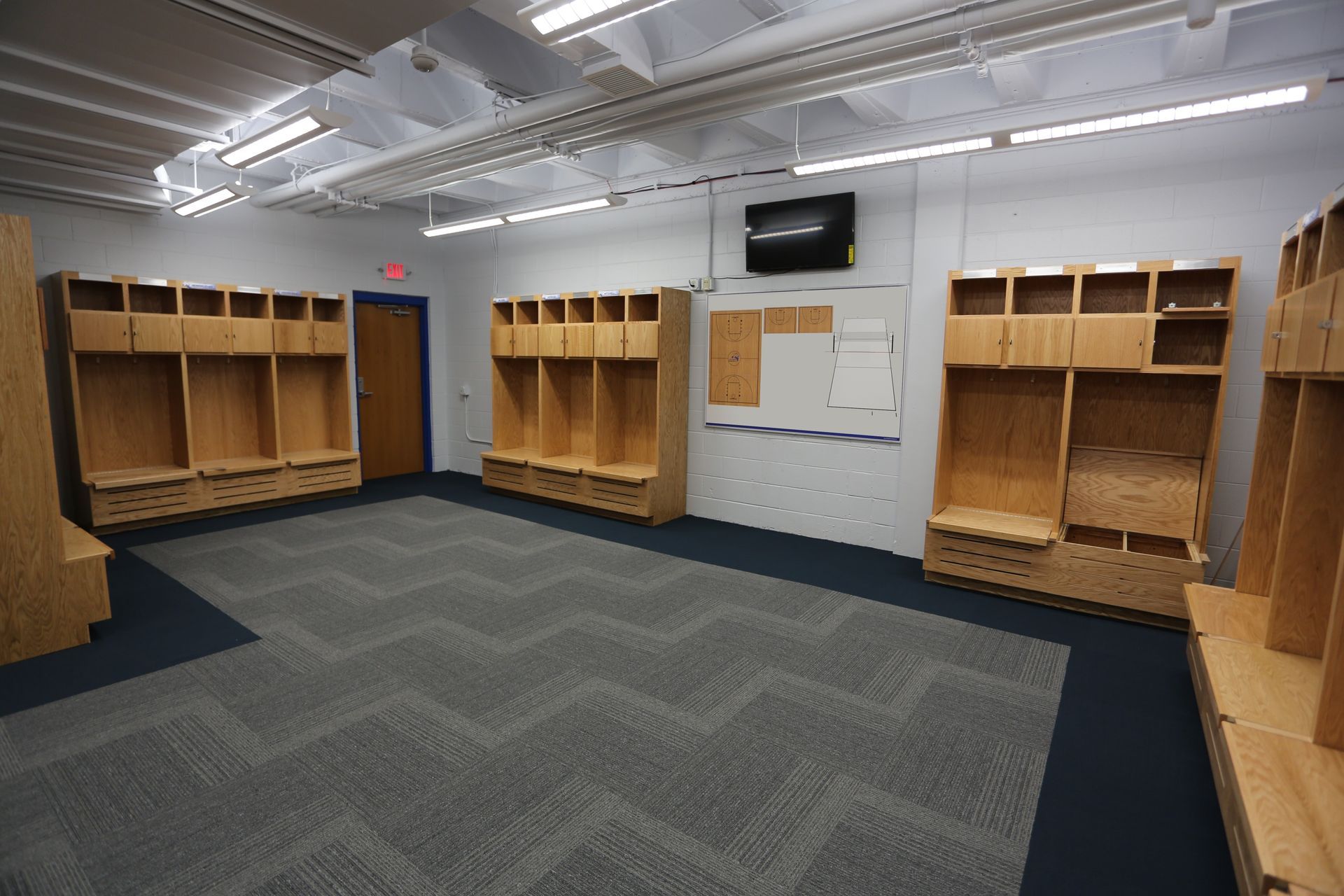 Empty locker room with light wood cubicles, a patterned gray carpet, and a wall-mounted whiteboard.