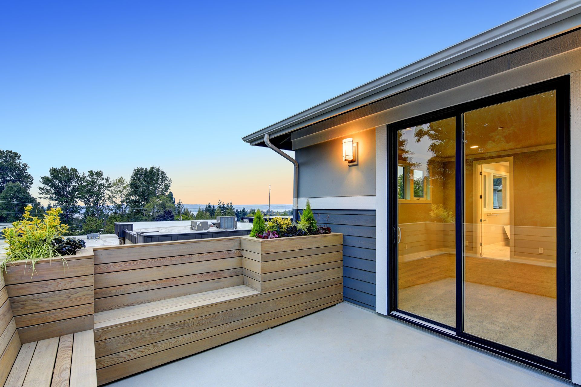A modern balcony featuring a built-in wooden bench, a large sliding glass door, and a sunset view of trees and water.