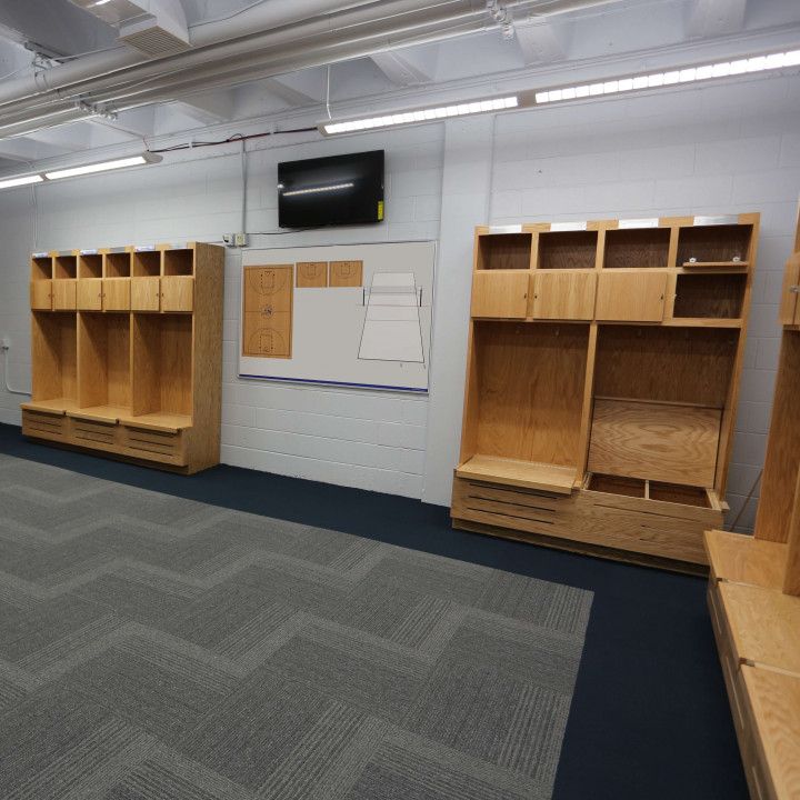 A locker room featuring wooden lockers, a white dry-erase board, a mounted television, and gray zigzag-patterned carpet.