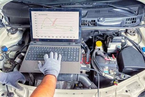 Laptop displaying engine data connected to a car engine. Mechanic's hands, in gloves, working.