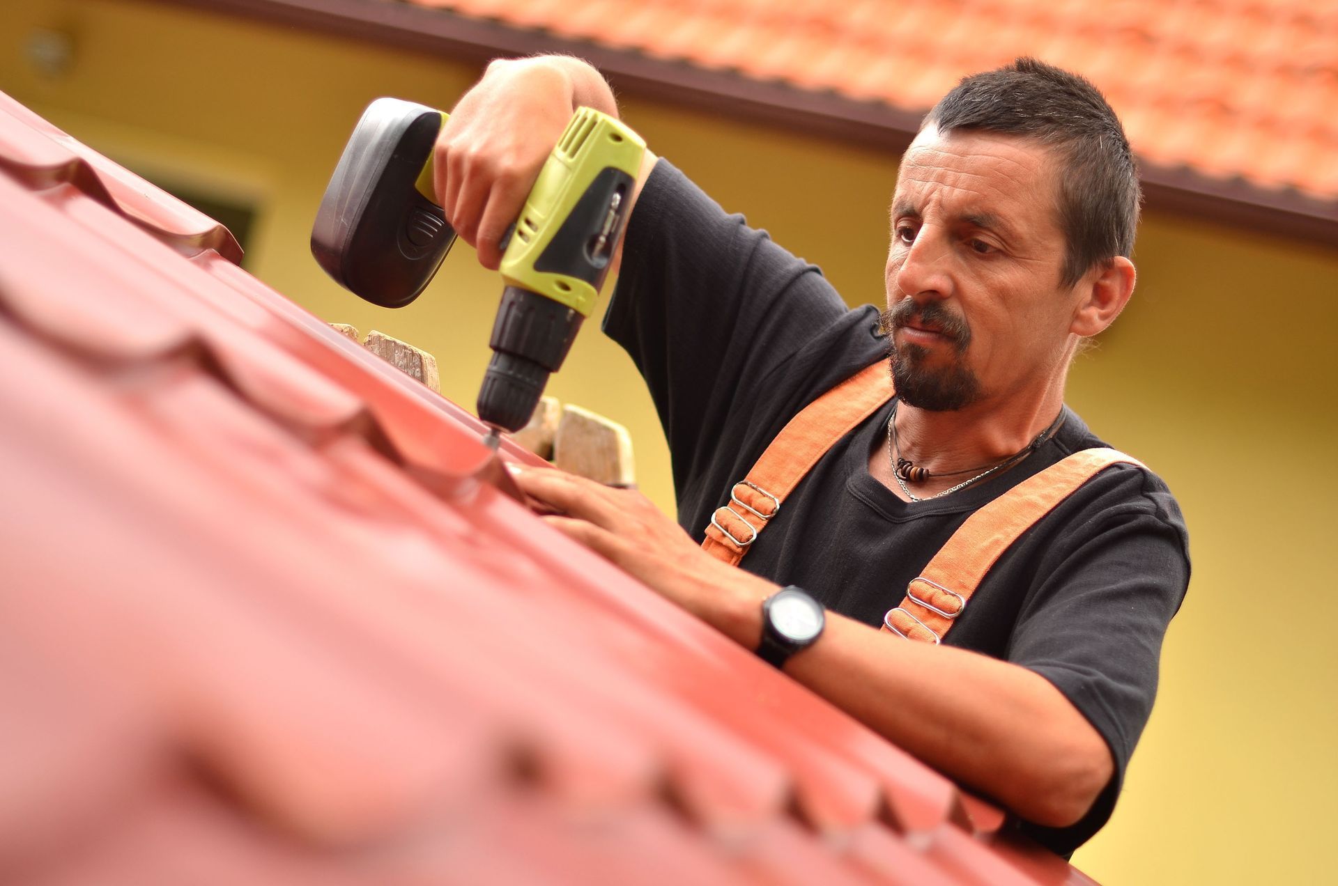 Man uses power drill on a red roof, wearing work overalls.