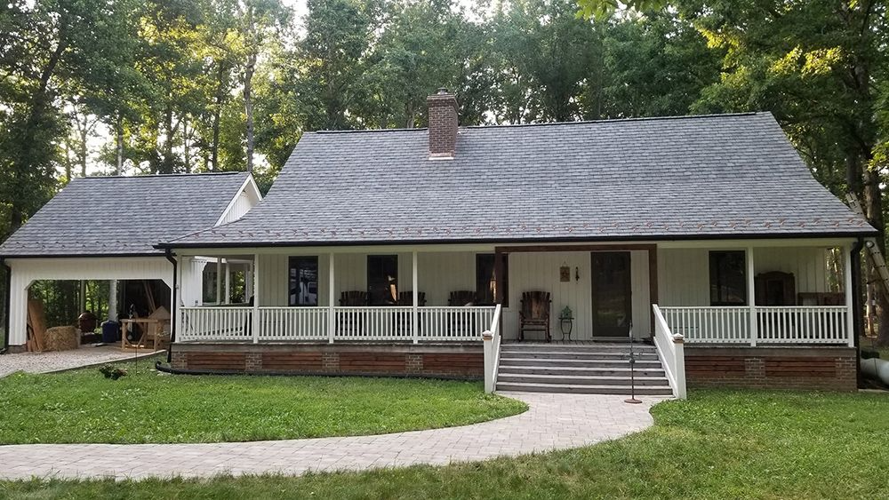 White farmhouse with porch, brick foundation, and gray shingle roof. A gravel path leads to the front steps.