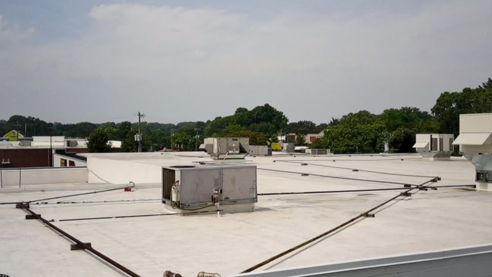 View of a flat commercial roof with HVAC units, wiring, and a distant treeline on a bright day.