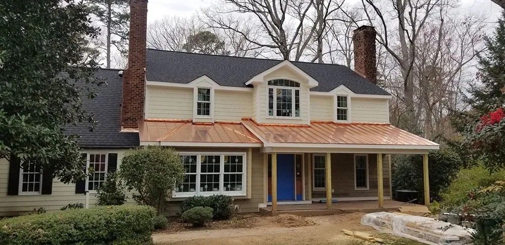 Two-story beige house with copper roof and blue door. Chimneys, windows, and porch are visible.