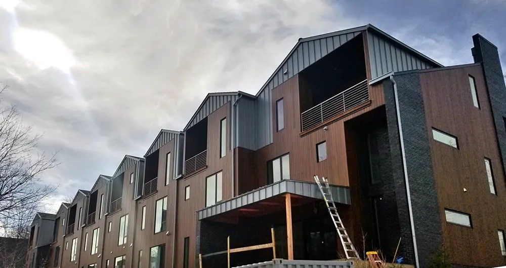Row of modern townhouses under a cloudy sky. Brown siding, grey metal roofs, balconies, and ladder.