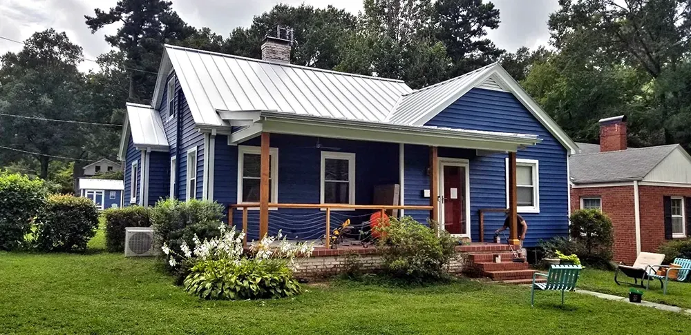 Blue house with white roof and porch, on a grassy lawn; another brick house in the background.