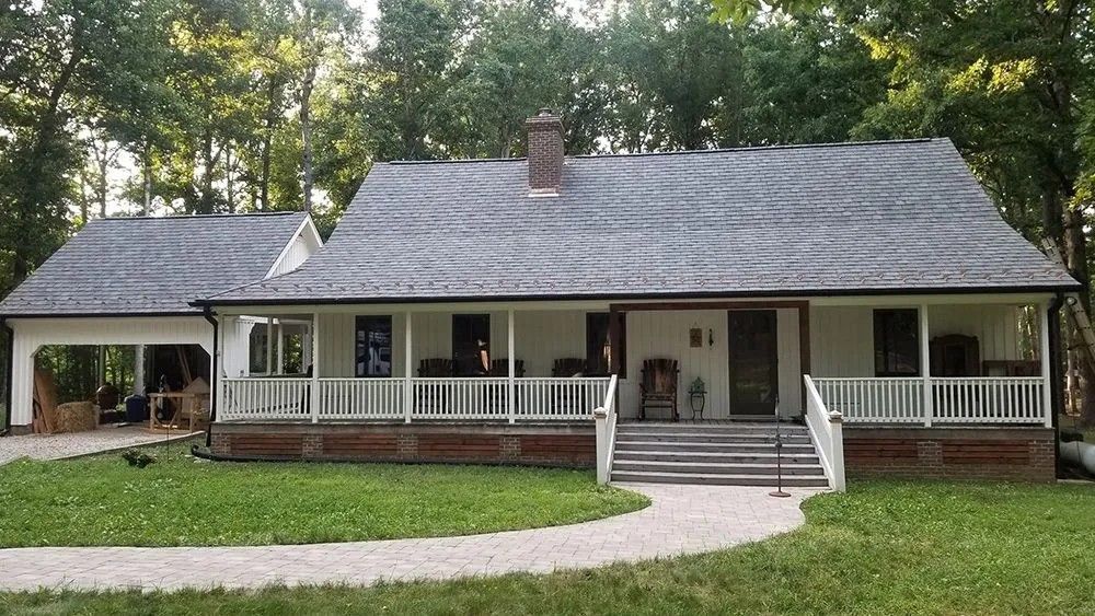 White farmhouse with porch and garage, gray roof, brick foundation, path leading to the front door, green lawn, trees in background.