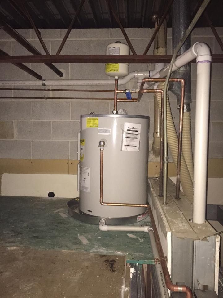 A water heater is sitting in a basement next to pipes.
