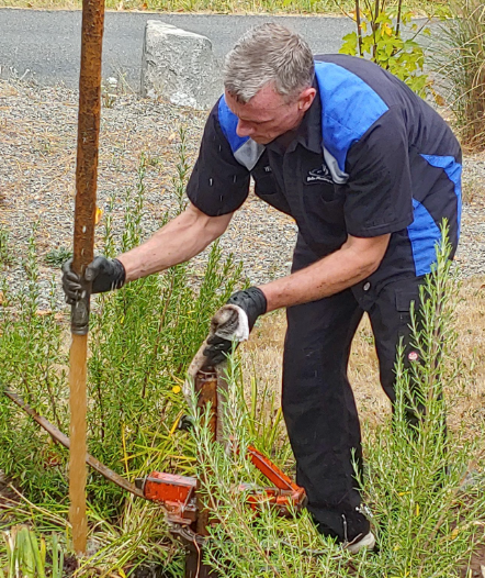 Man in blue and black shirt using a tool on a pipe in a grassy outdoor area.