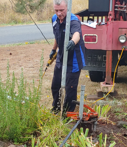Man working on a well, holding pipe. He's standing near a drilling rig, grass around him.