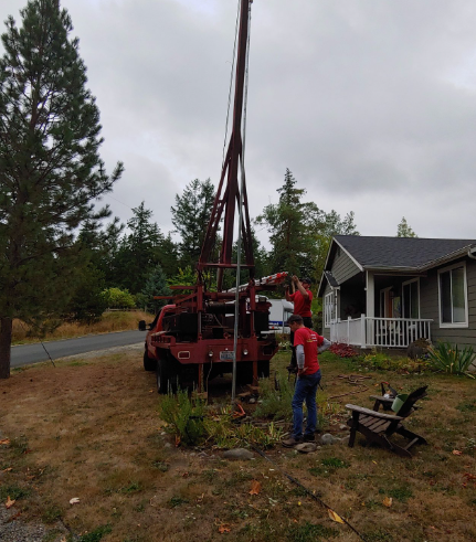 Red truck with drilling rig. Two people work near residential house, cloudy sky.