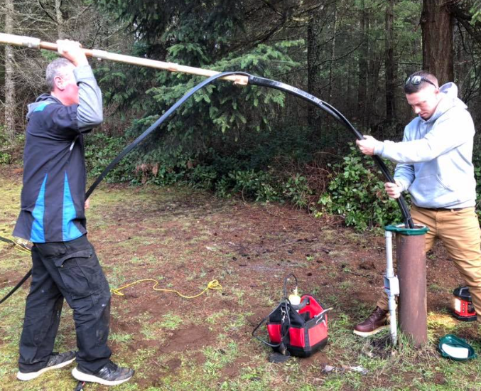 Two people manipulating a black hose near a well in a wooded area. One holds a pole.