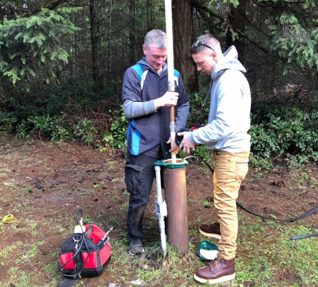 Two people working on a well in a wooded area. One holds pipe, the other adjusts parts.