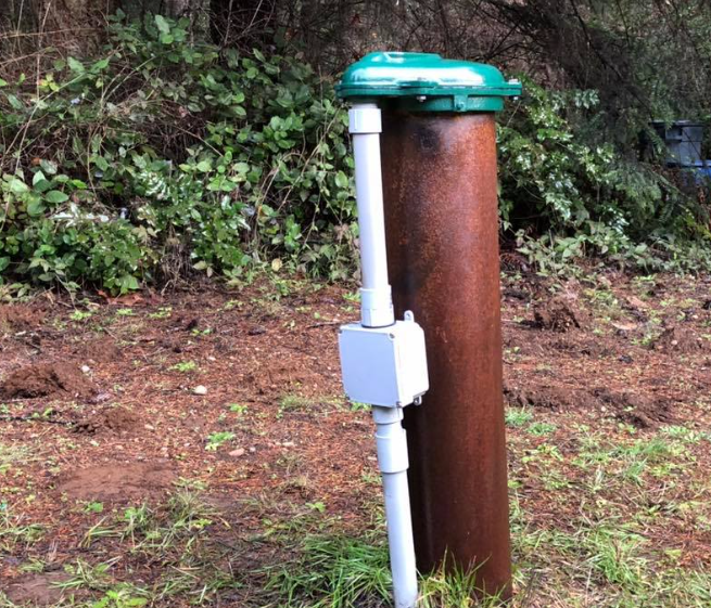 Rusty brown metal cylinder with green cap, white conduit, and electrical box in a grassy area.
